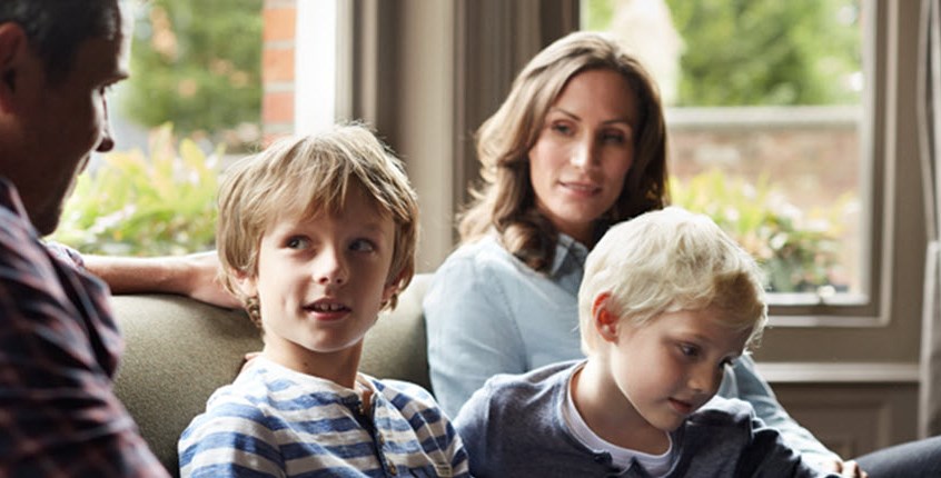 Mother, father, and two young children sitting together on the sofa, talking about chronic illness and how it will affect the family's future