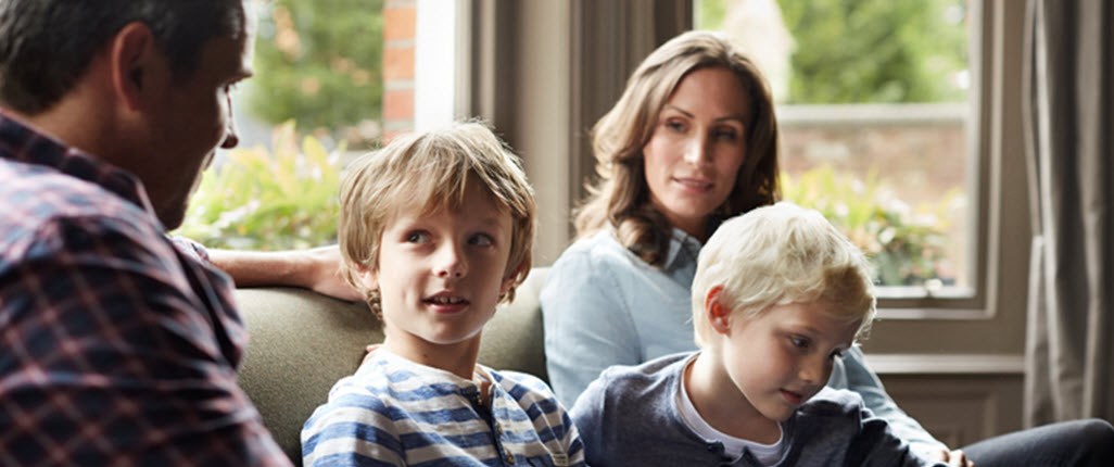 Mother, father, and two young children sitting together on the sofa, talking about chronic illness and how it will affect the family's future