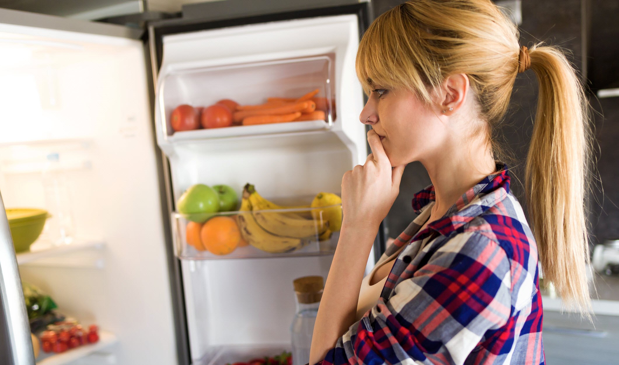 Woman staring thoughtfully into her fridge, unsure about her appetite