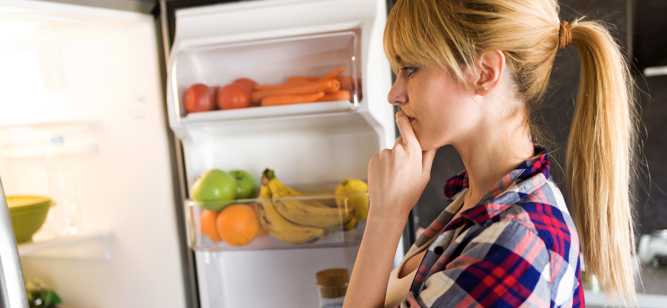 Woman staring thoughtfully into her fridge, unsure about her appetite