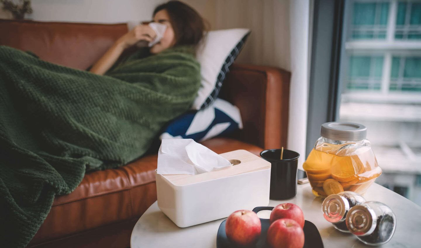 Woman with flu lying on her couch and blowing her nose with a tissue