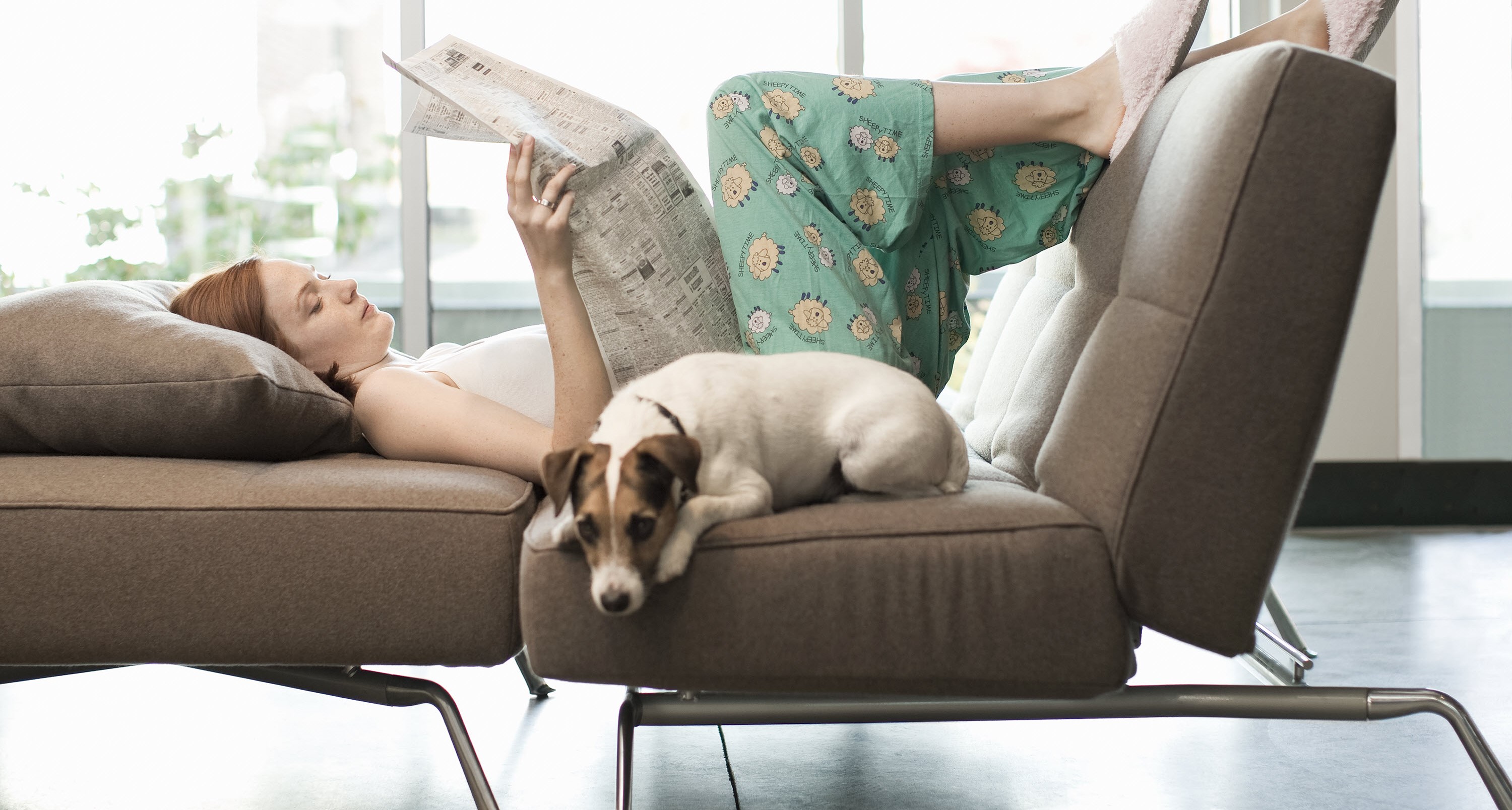 Woman relaxes on sofa with newspaper and dog, preserving her energy in case of migraine